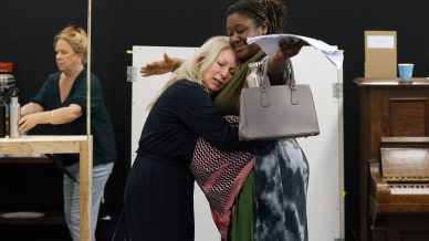 Two women embrace warmly; one clutches a script and a handbag while spreading her arms, the other leans her head on her shoulder. In the background, a third woman works by a table beside an upright piano.
