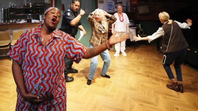 Five adults stand in a room with wooden floors; one woman in a colourful dress gestures passionately towards the camera, while others, including a man in a police uniform, react dramatically behind her, suggesting a lively performance.