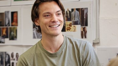 A young man with light brown hair sits on a pink chair, smiling and talking. He wears a light green T-shirt and blue shorts. Behind him are mood boards with photos and images on the wall.