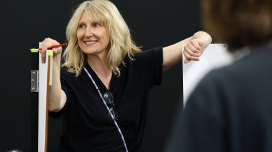 A smiling woman with blonde hair holds a pencil and leans against a board, wearing a black shirt and sunglasses on a strap around her neck, in a brightly lit indoor setting. Another person is blurred in the foreground.