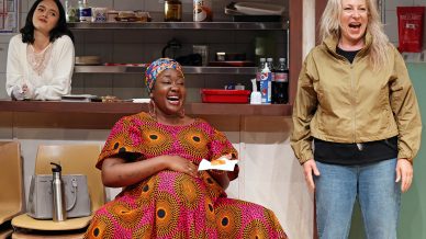 Three women are in a kitchen. One stands behind a counter, another sits and laughs holding food, wearing a colourful dress, and the third stands smiling, wearing a beige jacket and jeans. There are shelves with kitchen items in the background.