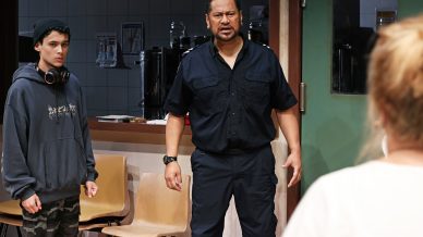 A man in a navy uniform stands in a room looking serious, whilst a young person in a hoodie and beanie with headphones round their neck stands nearby. Another person is partially visible in the foreground.