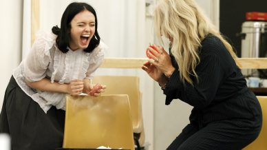 Two women laughing enthusiastically in a room; one stands behind a chair wearing a white blouse and black skirt, while the other, with long blonde hair, crouches in dark clothes, both appearing joyful and animated.