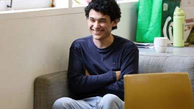 A young man with curly hair, wearing a navy blue long-sleeved shirt and jeans, sits on a grey sofa smiling with arms folded. There is a green shopping bag, a cup, and a green bottle on a table behind him.