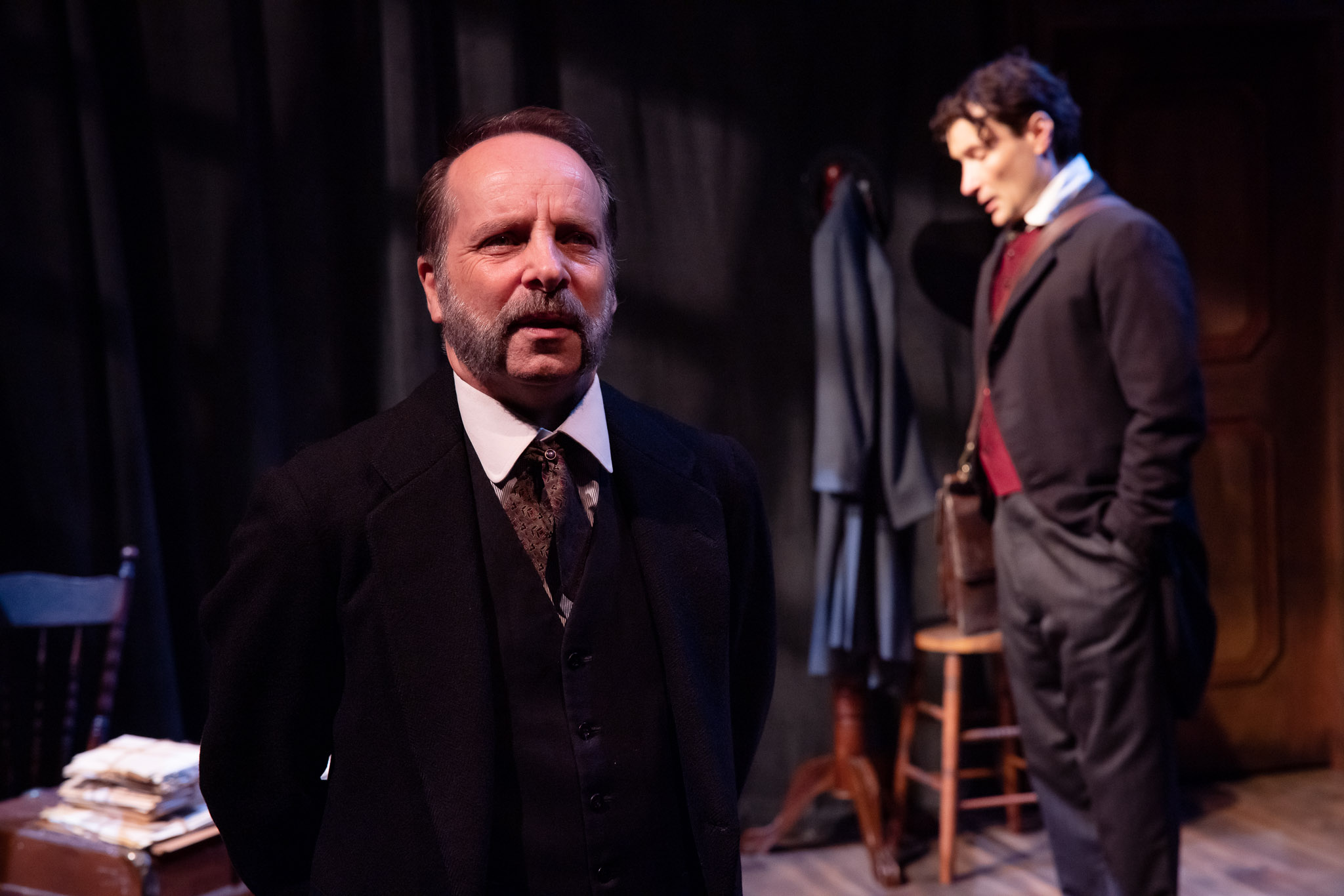 Two men in period suits are on a dimly lit stage. The older man in the foreground looks serious, whilst the younger man stands in the background near a coat stand, appearing contemplative.