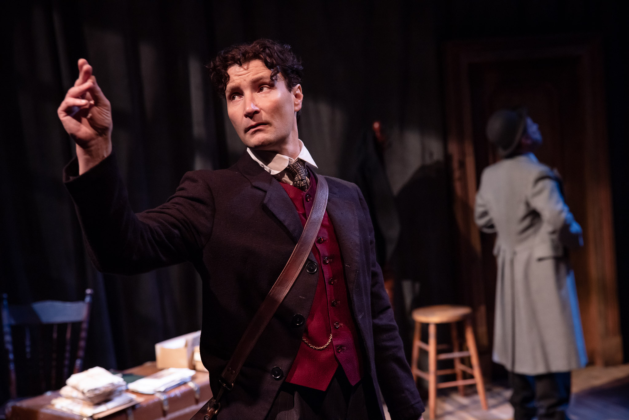 A man in a dark suit and red waistcoat stands with his hand raised, looking thoughtful. Another person in a coat and hat faces away near a wooden door. The scene appears to be set in a dimly lit study or office.