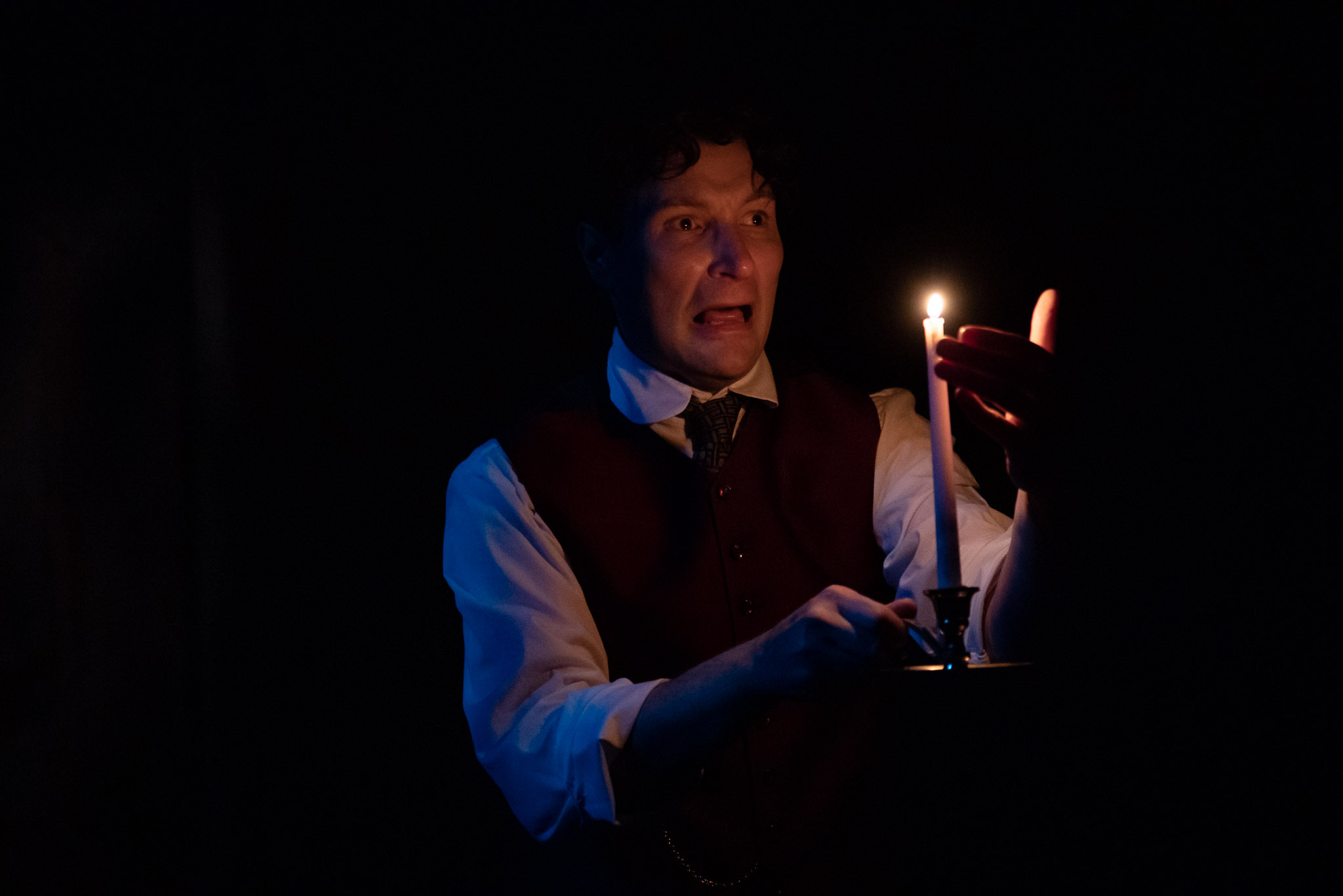 A man in a dark room holds a lit candle, wearing a waistcoat and tie; his face shows fear or shock as he looks towards the light.
