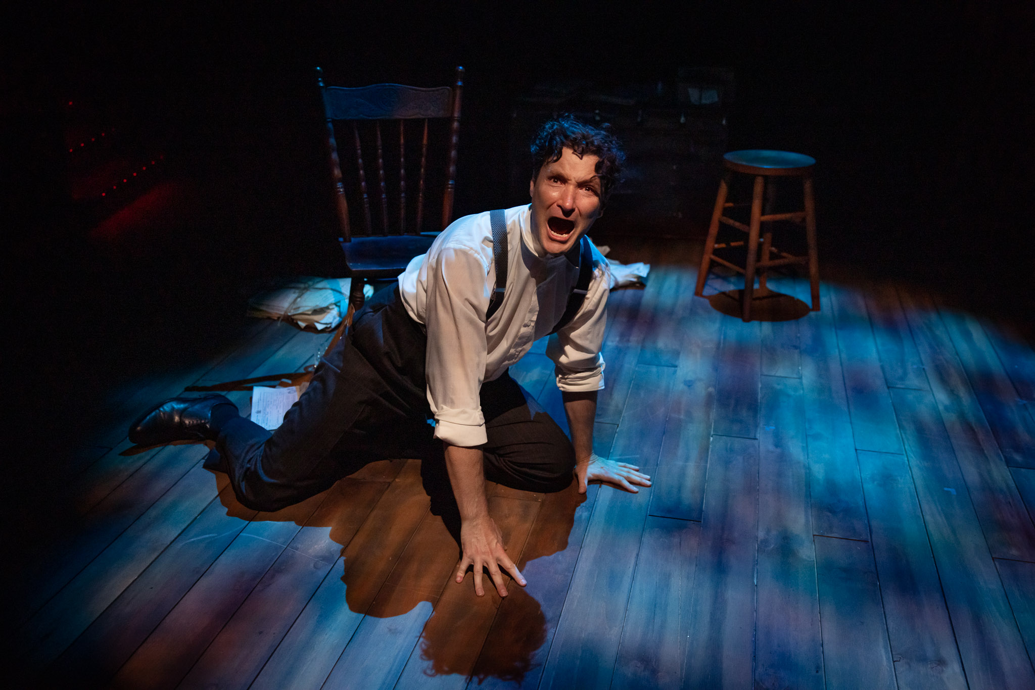 A man in dress trousers, a white shirt, and braces kneels on a wooden stage, looking distressed and shouting. A wooden chair and stool are in the background, with dramatic stage lighting highlighting the scene.
