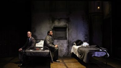Two men in suits sit back-to-back on separate beds in a dimly lit, worn-out room with peeling walls and minimal furnishings, creating a tense and dramatic atmosphere.