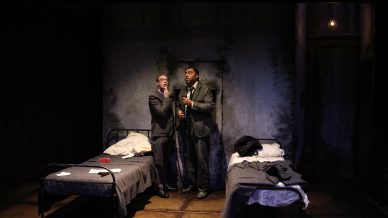 Two men in suits stand closely together between two single beds in a dimly lit, worn room with cracked walls. One man gestures towards his mouth while the other listens intently. Papers and coats are on the beds.