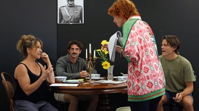 Four people rehearse a scene around a table set with flowers and candlesticks, while a black-and-white photo of a man in uniform hangs on the wall behind them. One woman stands holding scripts as the others sit and watch her.
