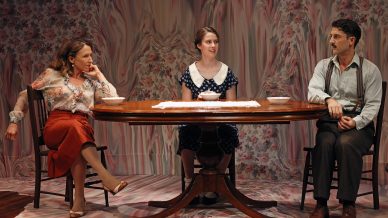 Three people sit around a wooden table with bowls in front of them, against a floral-patterned backdrop. The woman on the left looks at the others, the woman in the middle smiles, and the man on the right looks serious.