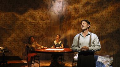 A man stands in the foreground speaking, while two women sit at a table in the background on a dimly lit stage. A large, smiling face is projected on the wall above them, creating a dramatic atmosphere.
