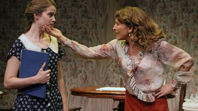 An older woman extends her hand towards a younger woman's face, who looks uneasy and holds a large book. They stand in a room with floral wallpaper and a wooden table in the background.
