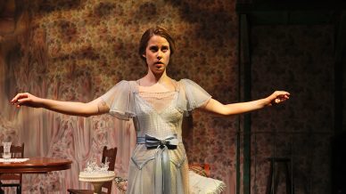 A young woman in a vintage blue dress stands with arms outstretched as another woman kneels, adjusting the hem. They are on a wooden stage with period furniture and floral wallpaper, creating a theatrical, old-fashioned setting.