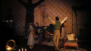 A man stands with arms raised, casting a large shadow on a wallpapered wall with a portrait. A woman in a white dress looks on. The room features vintage furniture and a gramophone, evoking a nostalgic atmosphere.