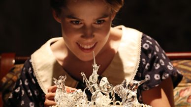 A young woman in a polka-dot dress smiles as she looks closely at delicate glass animal figurines arranged on a decorative cake stand.