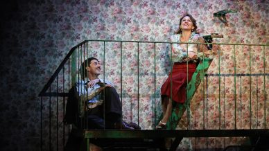 A man sits on the steps of a green fire escape, smiling up at a woman seated higher up. The background is floral wallpaper. Both appear to be engaged in cheerful conversation.