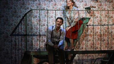 A man and woman sit on a metal fire escape against a wall with floral wallpaper. Both appear thoughtful; the man is seated on the railing, while the woman stands leaning beside him, mid-conversation. The scene is warmly lit.