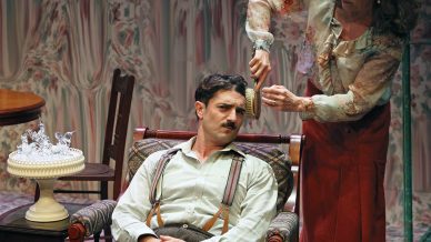 A woman in a floral blouse and red skirt stands behind a seated man, holding a comb to his hair. The man sits in an armchair, looking unimpressed. A table with a decorative cake is beside them, and the room has vintage décor.