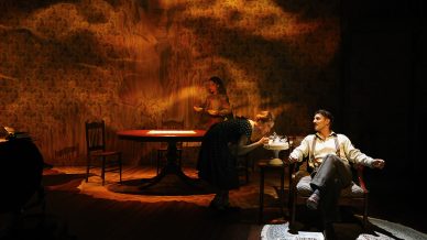 A woman leans over to blow out candles on a birthday cake held by a seated man at a dining table, while another woman stands behind them. Warm, dramatic stage lighting creates a nostalgic and intimate atmosphere.