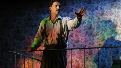A man with a moustache, wearing braces and a button-up shirt, stands on a balcony with one hand raised, appearing emotional. He is lit by colourful stage lights against a floral wallpaper background.