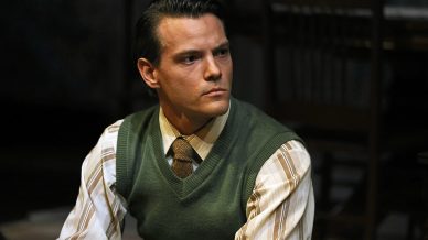 A man with neatly combed dark hair, wearing a green jumper vest over a striped shirt and brown tie, sits indoors looking to the side with a serious expression. Wooden chairs and a dim background are visible behind him.