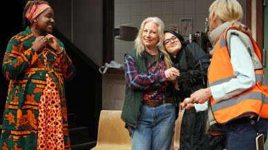 Four women stand together in a brightly lit room. One wears a colourful, patterned dress and headwrap, another hugs a smiling girl, and the fourth woman wears a reflective safety vest and holds a clipboard. They appear to be having a friendly conversation.
