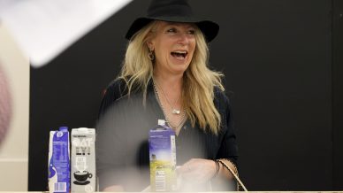 A woman with long blonde hair wearing a black hat and dark shirt smiles whilst standing behind a wooden counter with cartons of oat milk and other beverages in front of her.