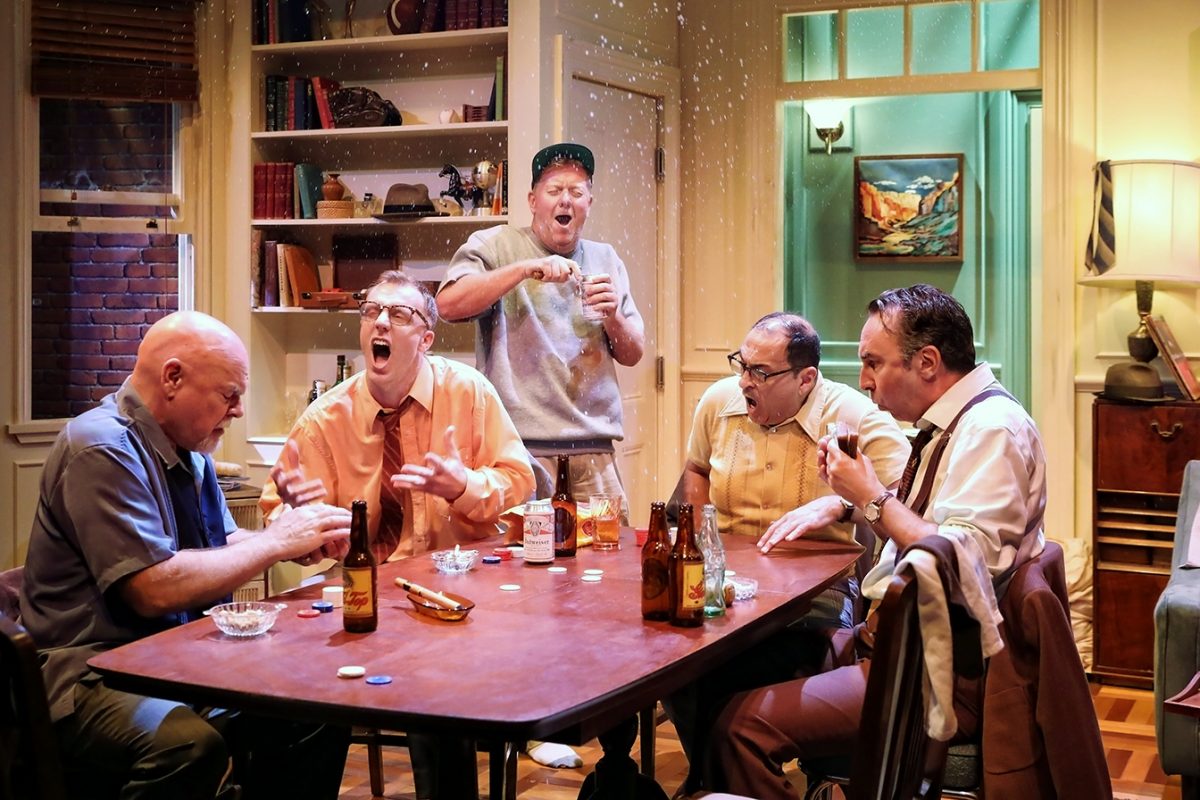 Five men sit around a table playing cards in a cosy, book-filled room. They react with surprise and laughter as one man in the background sprays liquid, capturing a lively, celebratory moment. Bottles and poker chips are on the table.