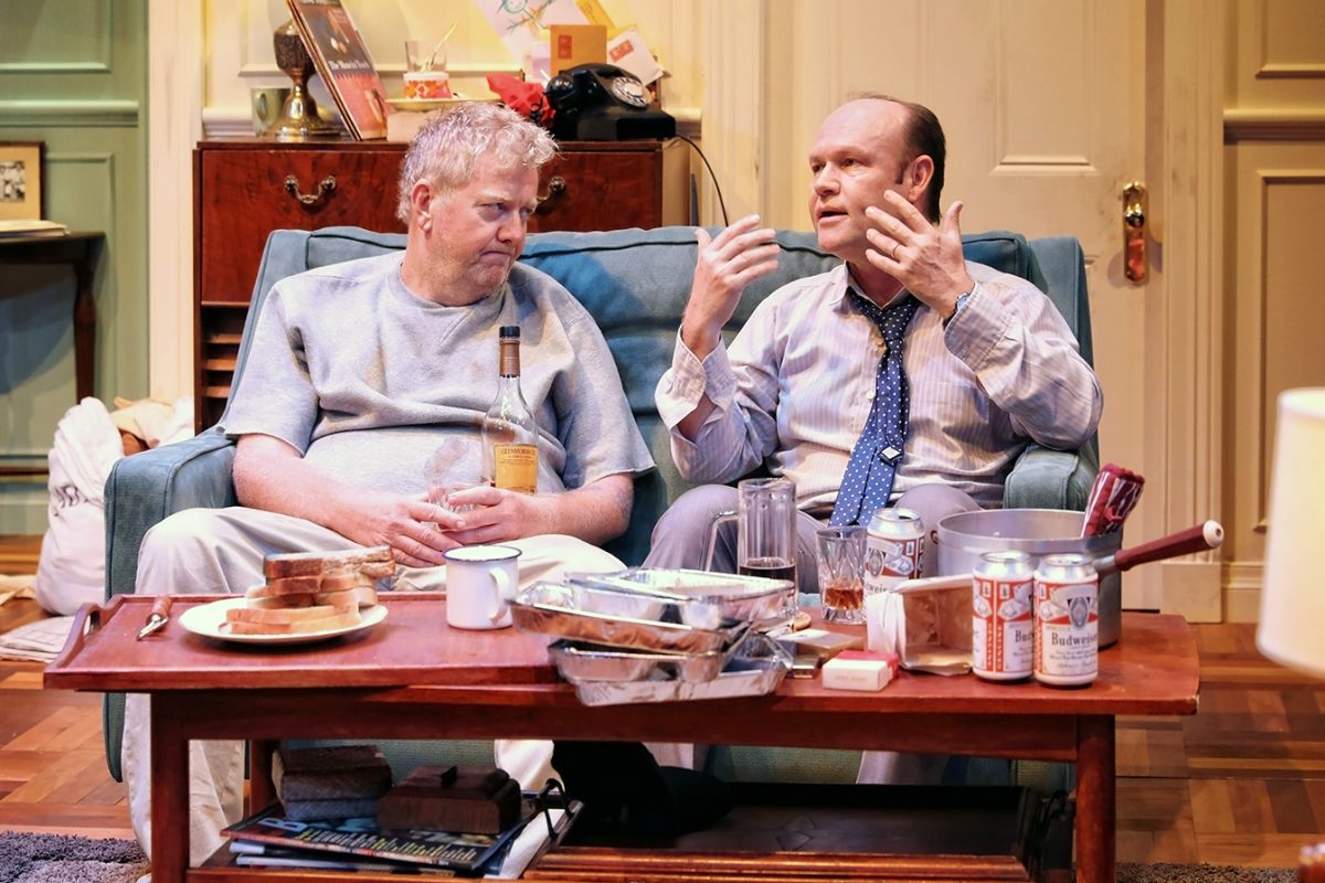 Two men sit on a sofa in a cluttered living room, with food, drinks, and beer cans on the coffee table. One man looks tired holding a bottle, while the other gestures animatedly during conversation.