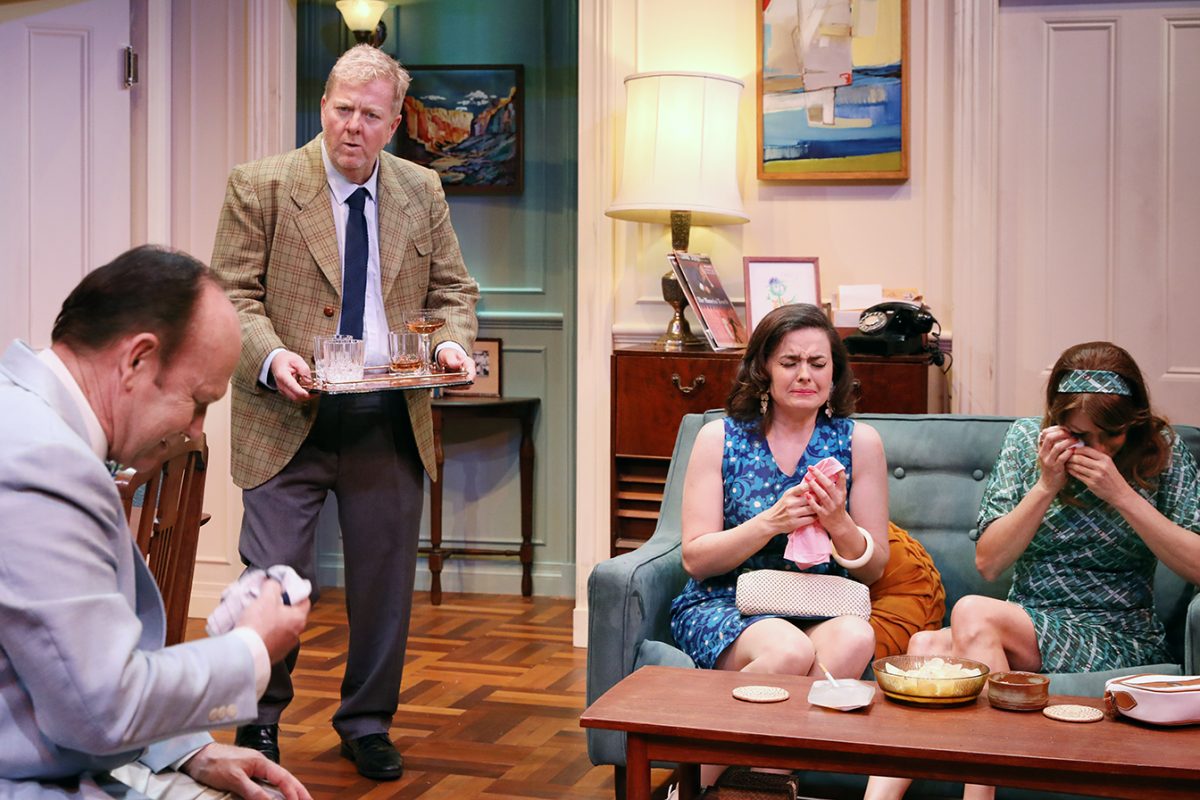 Four people sit in a living room set; two women on a sofa appear upset, wiping their eyes, while a man holds a tray with glasses and another man sits nearby, dabbing his face with a cloth. The scene is tense and emotional.