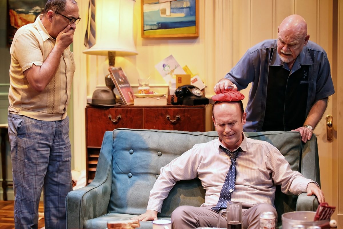 Three men in a living room; one man sits on a blue sofa with an ice pack on his head, placed there by another man, while the third man stands nearby, looking concerned with his hand covering his mouth.