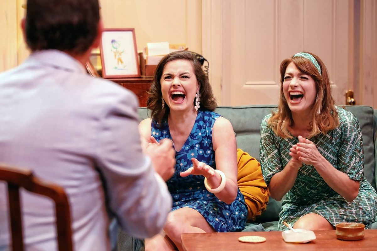 Two women in colourful dresses sit on a sofa, laughing and clapping excitedly while facing a man whose back is to the camera. The scene appears joyful and lively in a cosy living room setting.