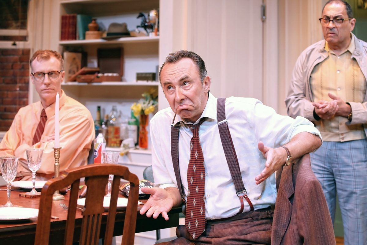 Three men in vintage clothing are in a dining room. The man in the centre gestures with a confused expression, while the other two men look on. The table is set for a meal, and shelves and cupboards are in the background.
