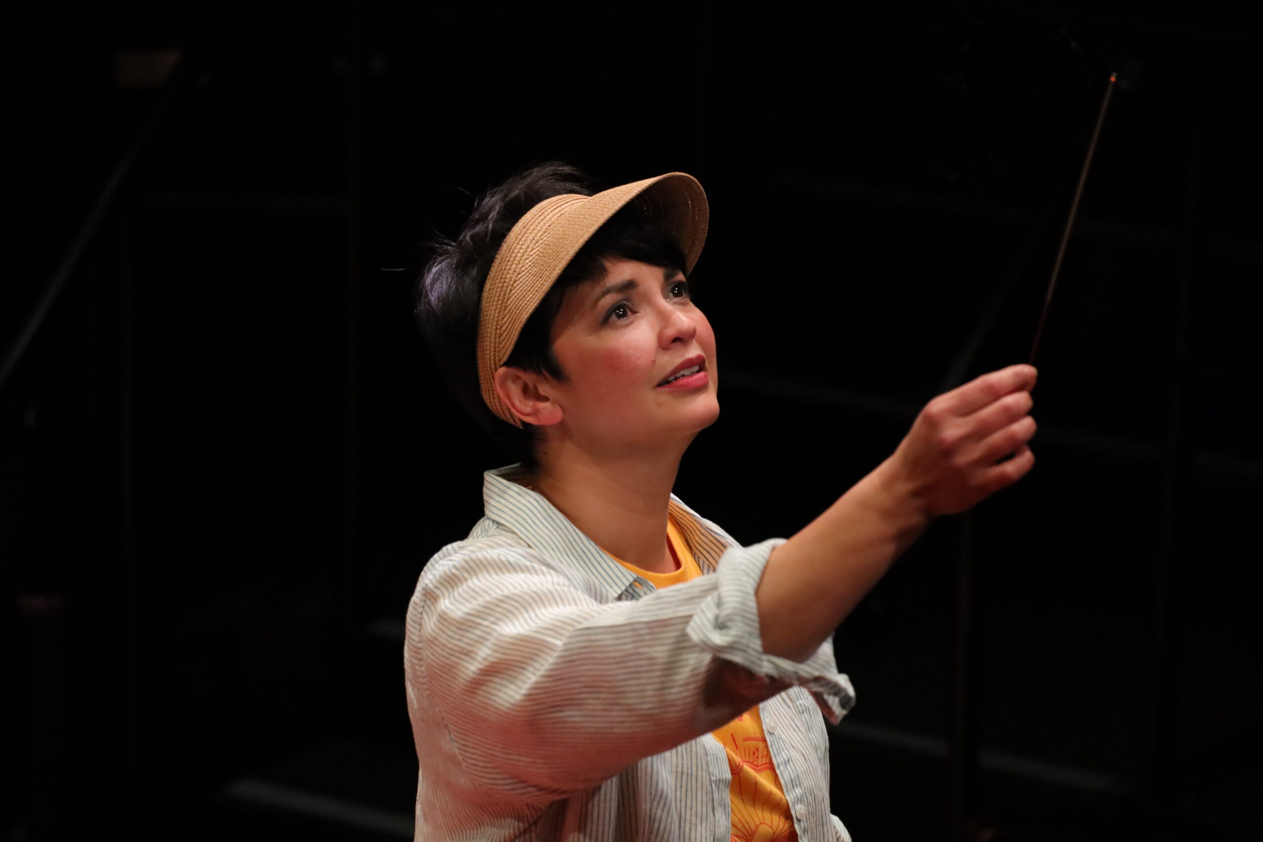 A person wearing a tan visor and a light striped shirt reaches upwards with a stick, gazing intently at something above them against a dark background.