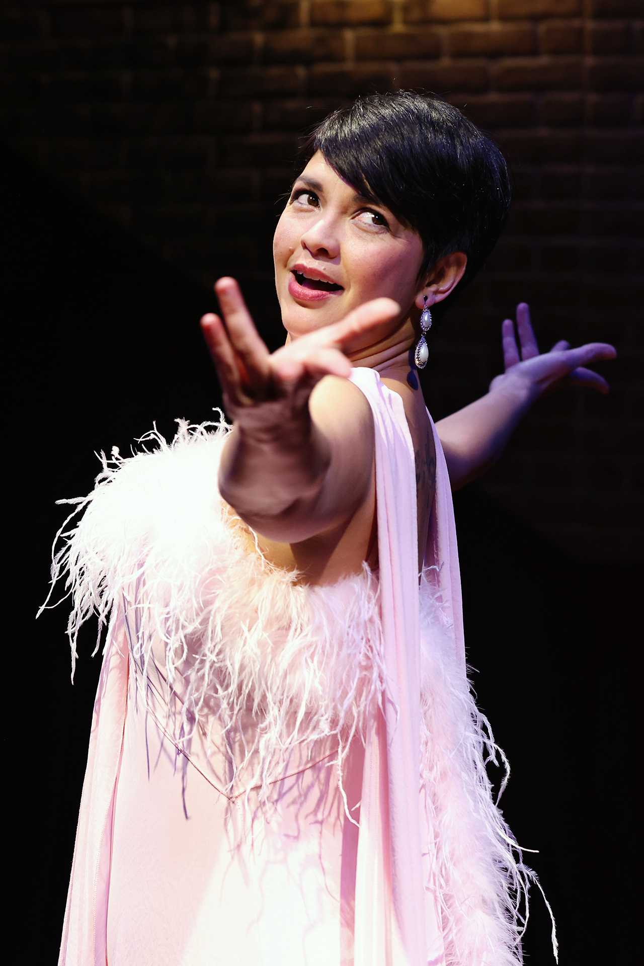A woman in a glamorous pink feathered dress performs on stage, extending one arm dramatically forward. She has short dark hair, wears dangling earrings, and smiles under theatrical lighting with a dark brick wall in the background.