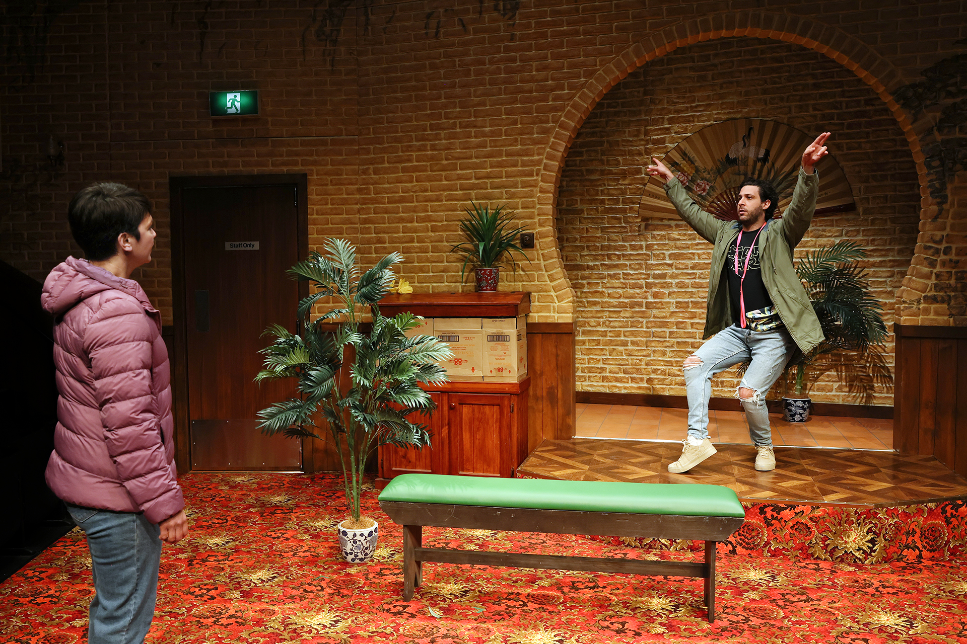 A person in a green jacket and ripped jeans enthusiastically gestures on a small stage, while another person in a pink puffer jacket stands watching. The set features faux brick walls, plants, and a red patterned carpet.