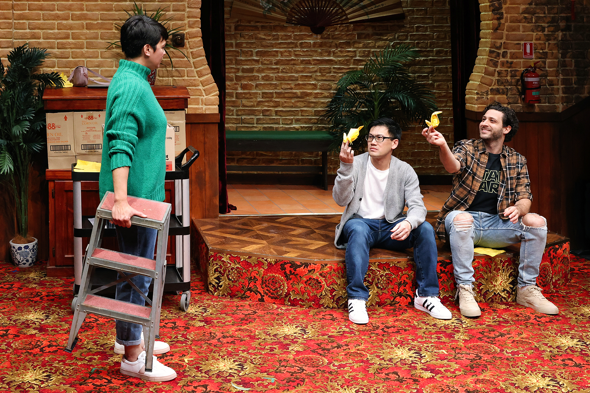 Three people in a room with red-patterned carpet and brick walls; one stands holding a step stool, while two sit on the floor, smiling and holding yellow origami birds. The setting appears playful and casual.