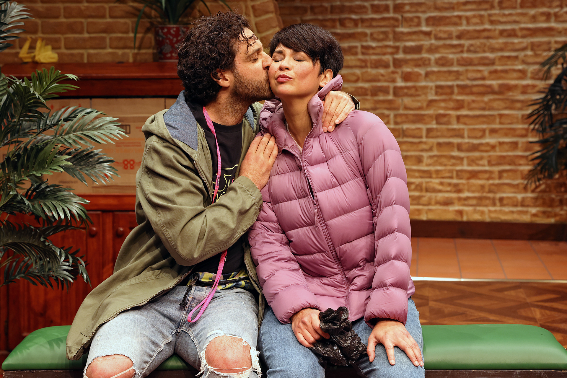A man kisses a woman on the cheek as they sit on a bench indoors. The man wears a green jacket and ripped jeans, whilst the woman, in a pink puffer jacket, smiles with her eyes closed. Green plants and a brick wall are in the background.