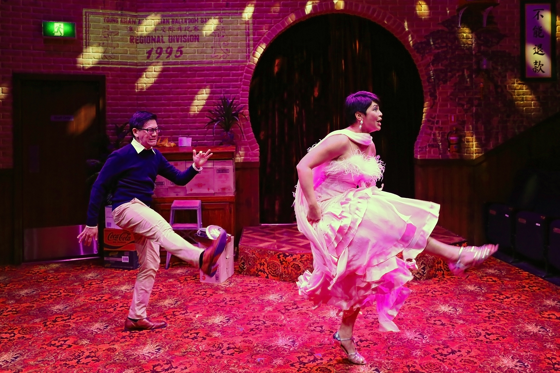 A man in glasses and a woman in a pink dress joyfully dance together, kicking their legs in sync on a vibrant red-carpeted stage with brick walls and colourful lighting.