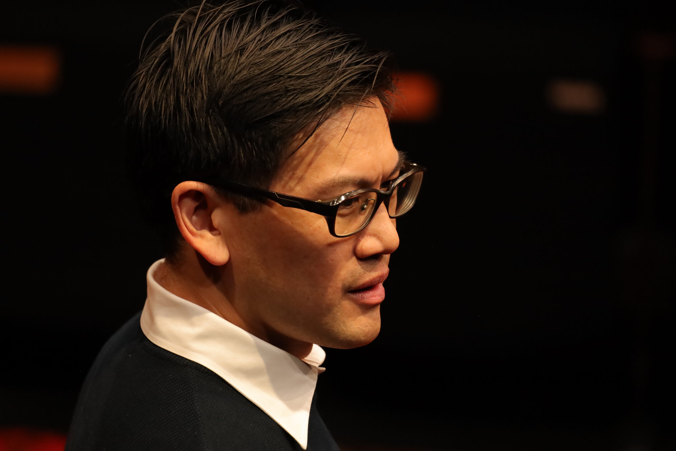 A man with short dark hair and glasses, wearing a white-collared shirt under a dark jumper, is facing left against a dark, blurred background.
