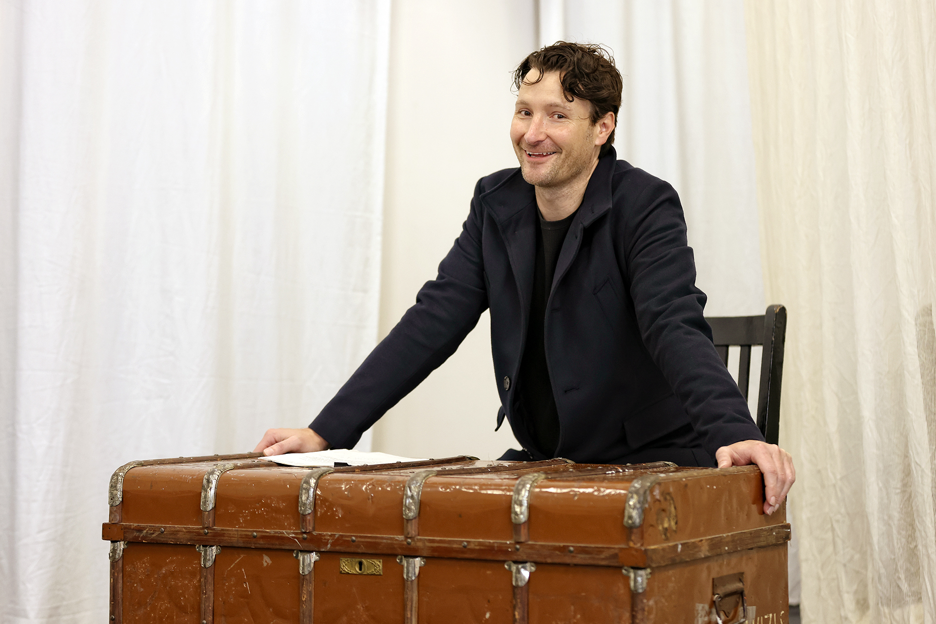 A man with curly hair and a dark jacket smiles whilst leaning on a large, old brown trunk. He is standing behind a wooden chair, with white curtains in the background.