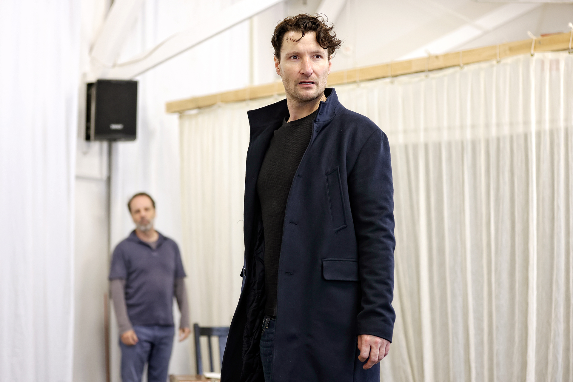 A man in a dark coat stands in the foreground, looking slightly to the side with a serious expression. Another man in casual clothes stands blurred in the background inside a brightly lit room with white walls and curtains.