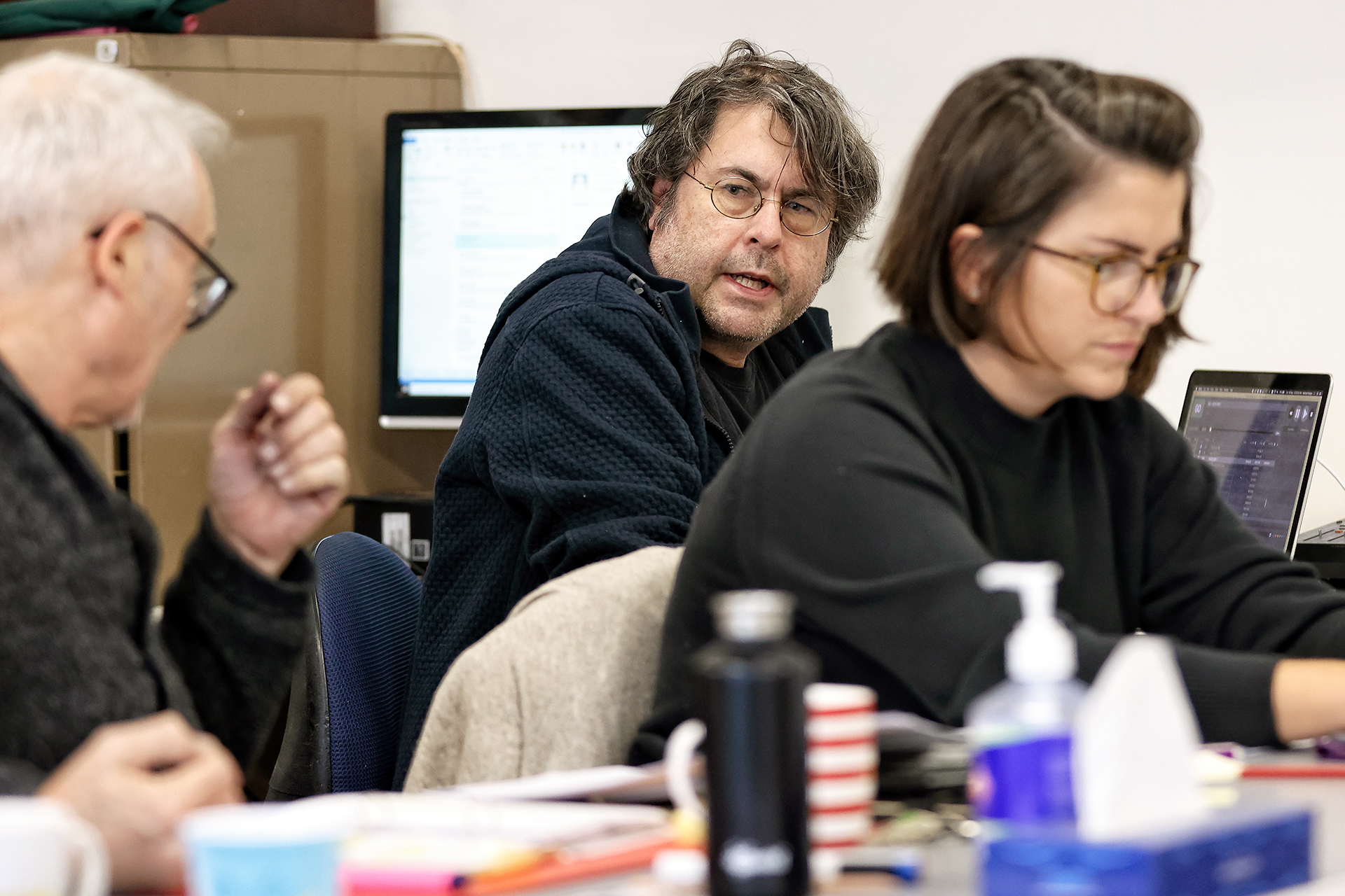 Three people are sitting at a desk with laptops, papers, and office supplies. The person in the middle with glasses and wavy hair looks towards the camera, while the others are focused on their work.