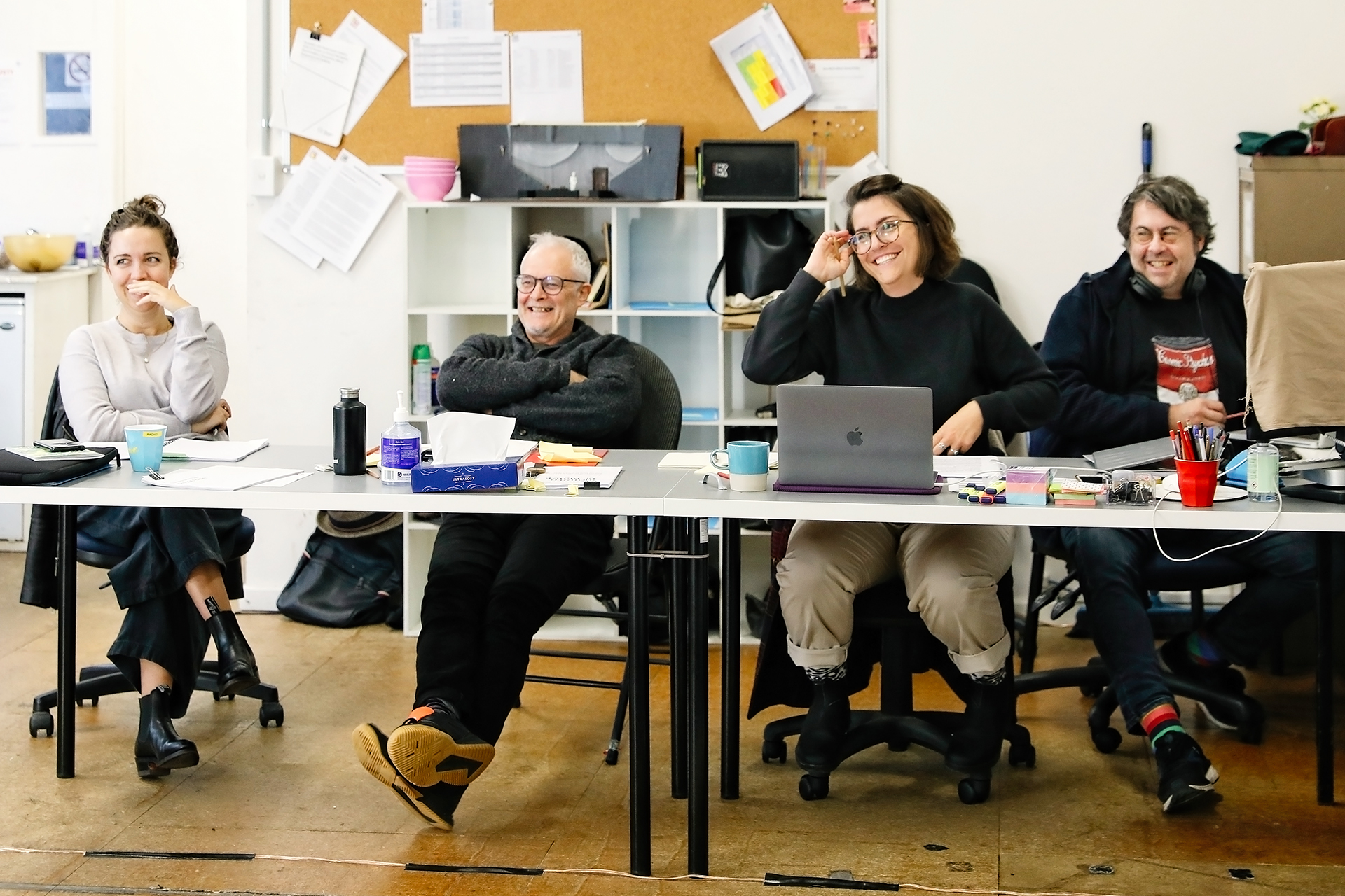 Four people sit at a long table in an office, smiling and appearing engaged. Laptops, papers, cups, and office supplies are on the table. Shelves and a noticeboard are visible in the background.
