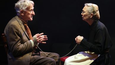 An elderly man and woman sit facing each other in dramatic lighting. The man’s wrists are bound with rope that connects to the woman’s hands. Both have serious expressions, suggesting a tense or emotional moment on stage.