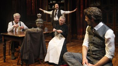 Four actors perform on a stage set resembling a vintage living room. Three sit—two knitting at a table, one in a waistcoat on a bench—while a standing man in the background gestures with arms outstretched.