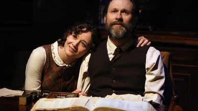 A woman smiles and rests her head on the shoulder of a bearded man as they sit together at a desk with open ledgers and papers, both wearing vintage-style clothing in warm lighting.