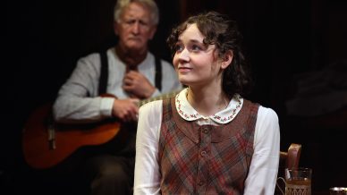 A young woman with short curly hair and a checked dress sits at a table, looking upward and smiling. An older man in the background, wearing braces, holds a guitar. A drink and a tray are on the table.
